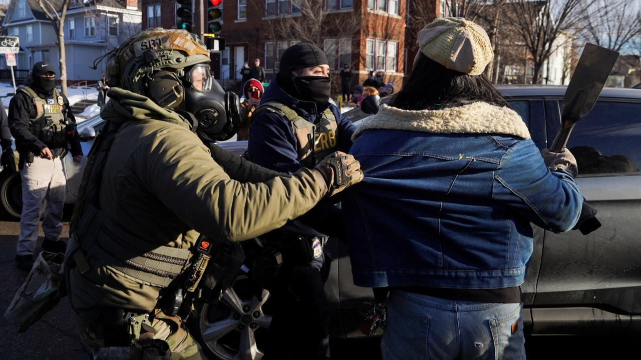 An agent in green uniform with a helmet and gas mask grabs a jacket worn by a protester on 13 January 2026. Other masked agents are nearby. There is snow on the ground on this bright winter's day.  