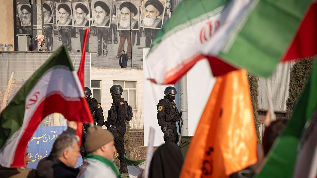 Iranske specijalne policijske snage u crnom pod punom opremom, sa maskama na licima, nadziru provladin miting u Teheran početkom meseca, na kojem ljudi nose zastave različitih boja, iznad policajaca su crnobele fotografije ajatolaha