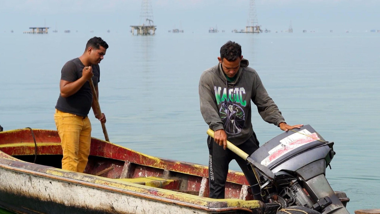 Dva mladića stoje u malom otvorenom čamcu na jezeru. Onaj levo, u žutim pantalonama i crnoj majici, drži drveni štap. Momak desno, u sivoj dukserici sa reči Magija ispisanom zelenom i belom bojom, drži ruke na sivom vanbrodskom motoru. Naftne platforme izdižu se iz vode u pozadini.