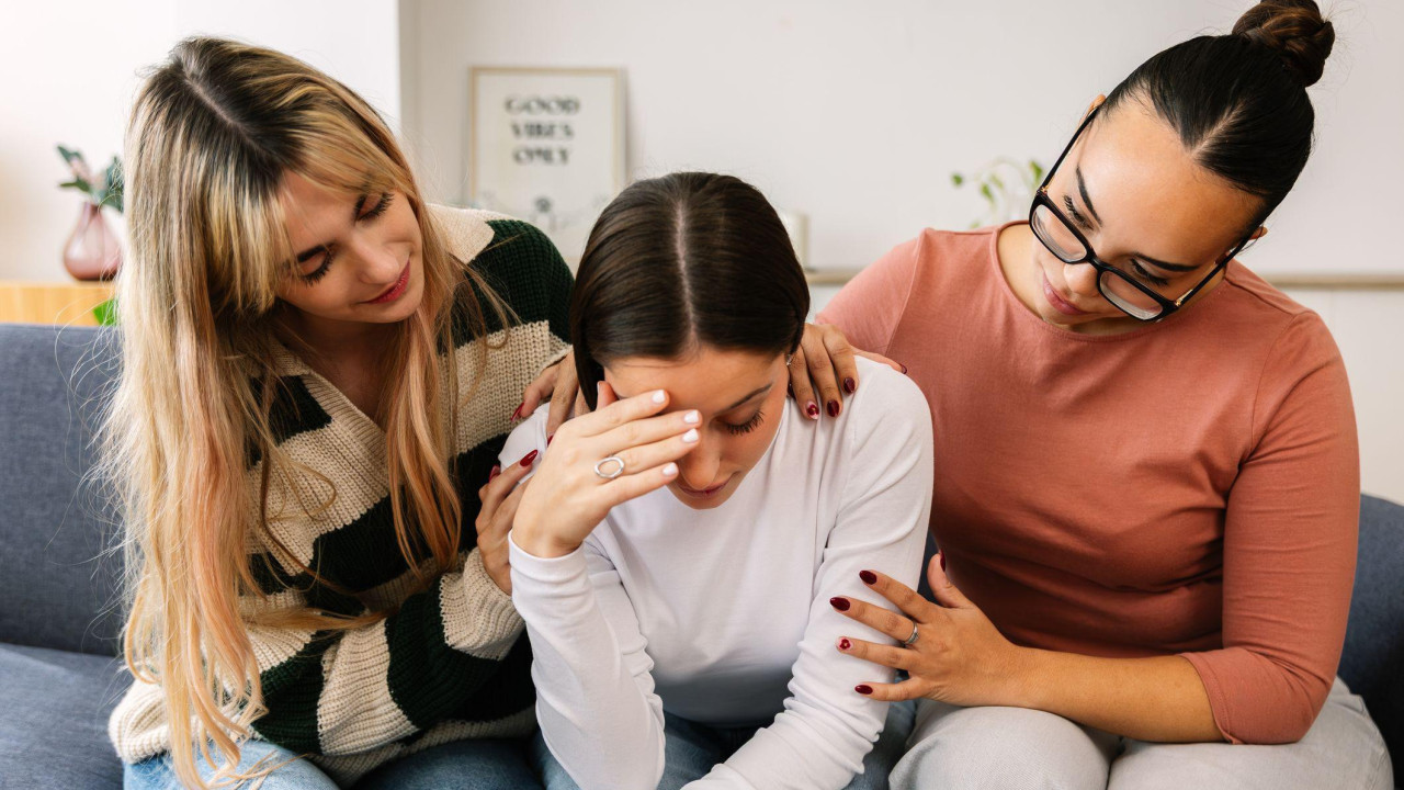 Dve mlade žene teše prijateljicu koja sedi između njih na sofi, sa rukom na licu i izgleda uznemireno.