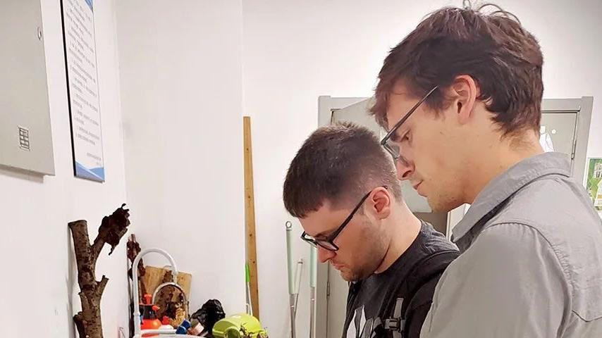 Dva muškarca, jedan u sivoj majici, a drugi u crnoj, gledaju u poslužavnik sa pečurkama u prostoriji sa belim zidovima