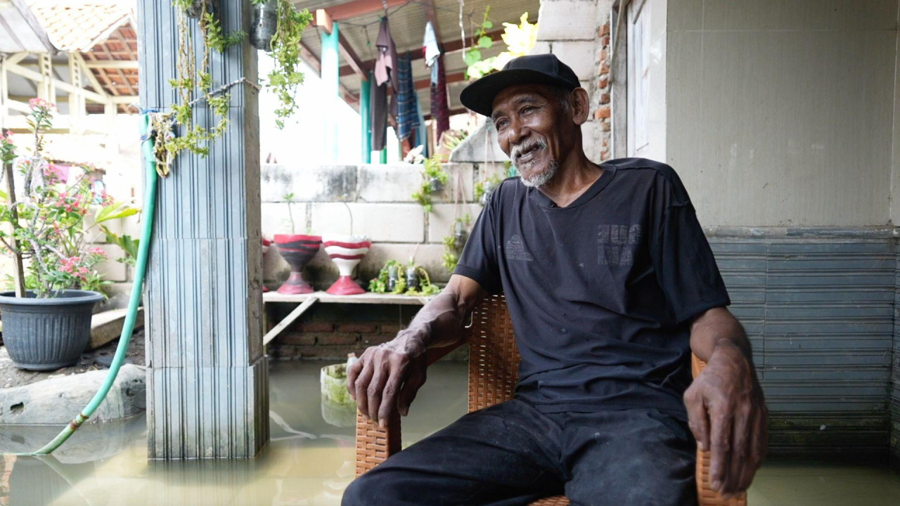 Suvandi sedi na stolici u crnoj majici kratkih rukava i pantalonama, ima crni kažčet na glavi i bradu, osmehuje se. Sedi u poplavljenoj kući