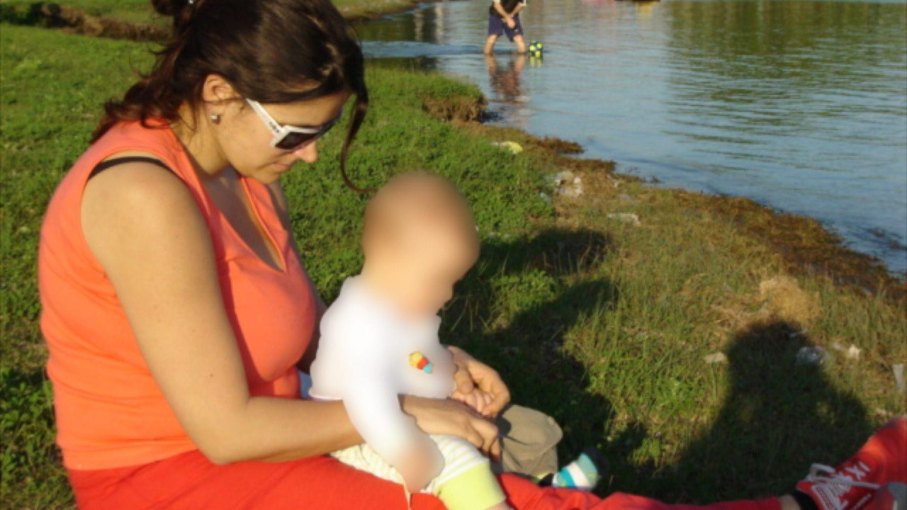 Rakel sedi na travi pored vode sa bebom, čije je lice zatamljeno, u krilu 