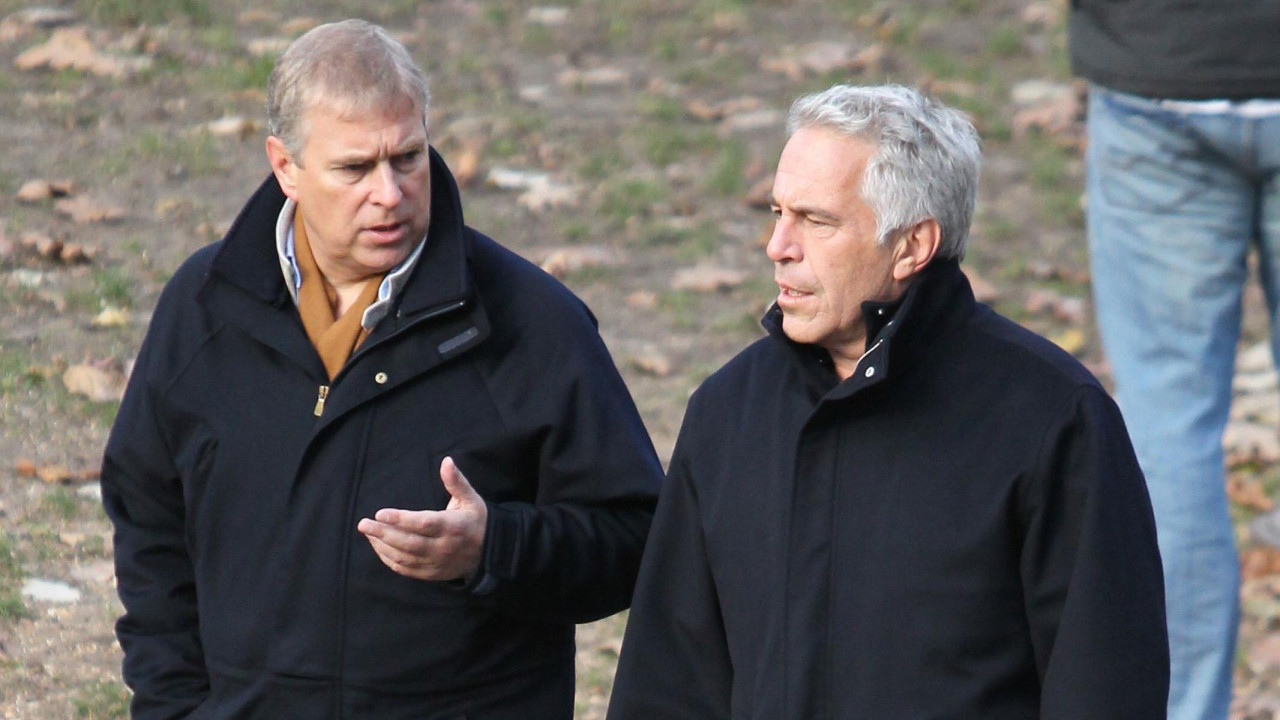 Andrew talks to Jeffrey Epstein as they walk. Both are wearing dark coats. Andrew's left hand is gesturing as he talks to Epstein. The ground behind them has grass and brown leaves on it. Someone with blue jeans is behind them and only their legs and waist are visible.
