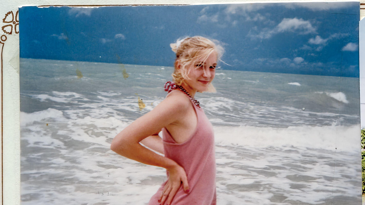 VIctoria Guiffre standing in the sea, wearing a pink dress with splashes on the lower part. Her hands are held to her hips. She is looking at the camera with hair tied back. The sea is grey and there are clouds in the sky. The photo is within a photo binder.