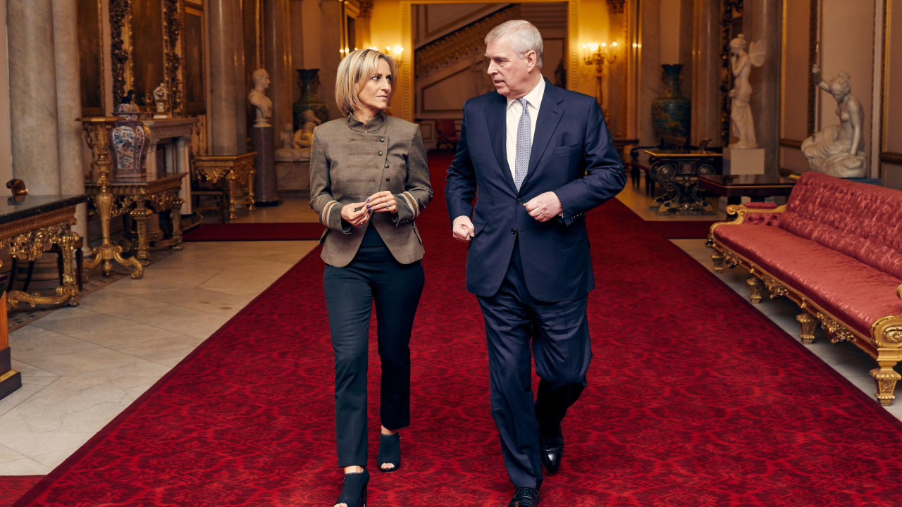 Emily Maitlis walking with Andrew through Buckingham Palace. They are on a red carpet with marble walls and statues. A long red seat is running down the side of the corridor.