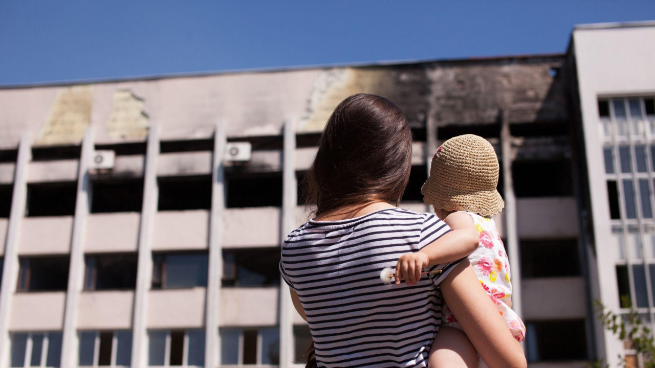 Majka sa bebom gleda na izgorelu zgradu gradske skupštine Marijupolja, okrenuta leđima kameri. Žena ima dugu tamnu kosu i nosi prugastu majicu, dok beba ima cvetnu haljinu i gole noge, sa šeširom za sunce. Zgrada ima ugljenisan deo na vrhu.