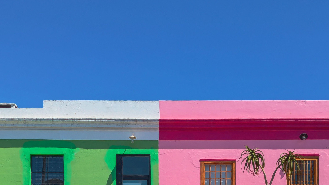 Pastelno plavi automobil - prazan putnika - stoji ispred jarko obojenih kuća, jedne zelene, jedne ružičaste, pod besprekorno plavim nebom.