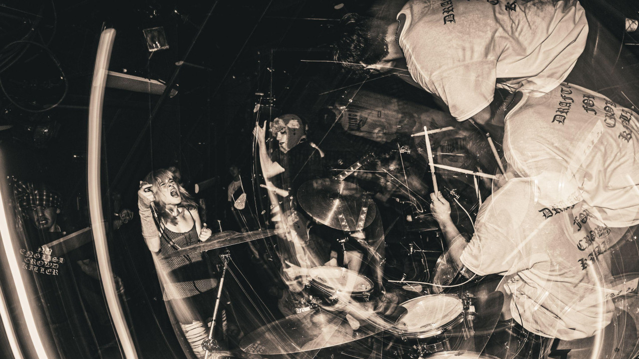 Fotografija zamućena pokretom prikazuje pevača, gitaristu i bubnjara pank benda Sailbout kako nastupaju na sceni.