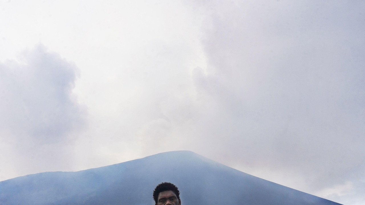 Bos na steni, obučen u srebrno odelo sa rukavicama i držeći odgovarajuću kacigu sa pričvršćenim vizirom, muškarac pozira za portret dok vulkan iza njega šalje oblak gasa i sumpora u nebo. 