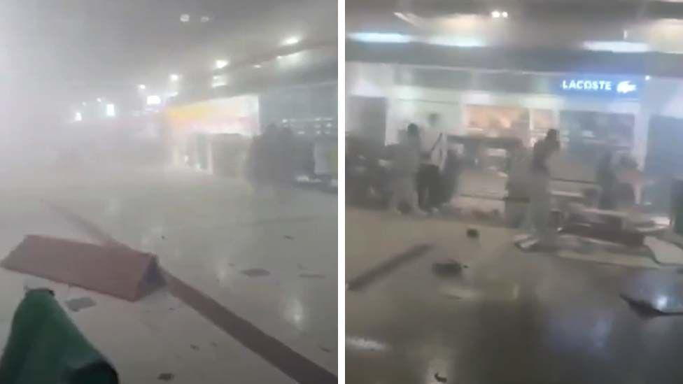 
                    A composite image with two frames showing the inside of Dubai airport filled with smoke, some figures can be seen walking through the smoke and debris on the floor
      