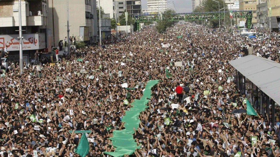 A wide city street packed with thousands of male and female protests on 15 June 2009. Some people are holding green flags, while others are holding posters. Tall buildings are on either side and a metal bridge is in the background.