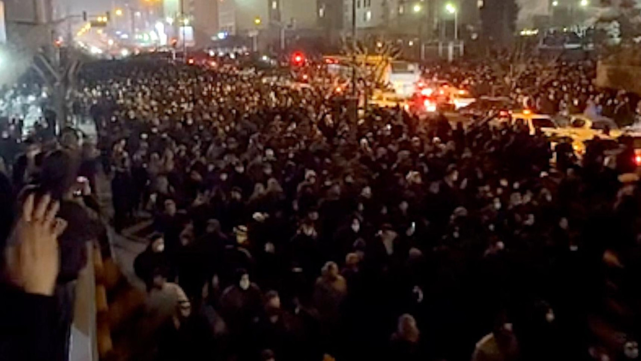 A view from a balcony at night of a street filled with people, stretching into the distance, marching towards the camera on 8 January 2026. The road is lit by streetlights, car headlights and lights from buildings. To the left is a balcony where people are watching. A woman nearby is clapping.
