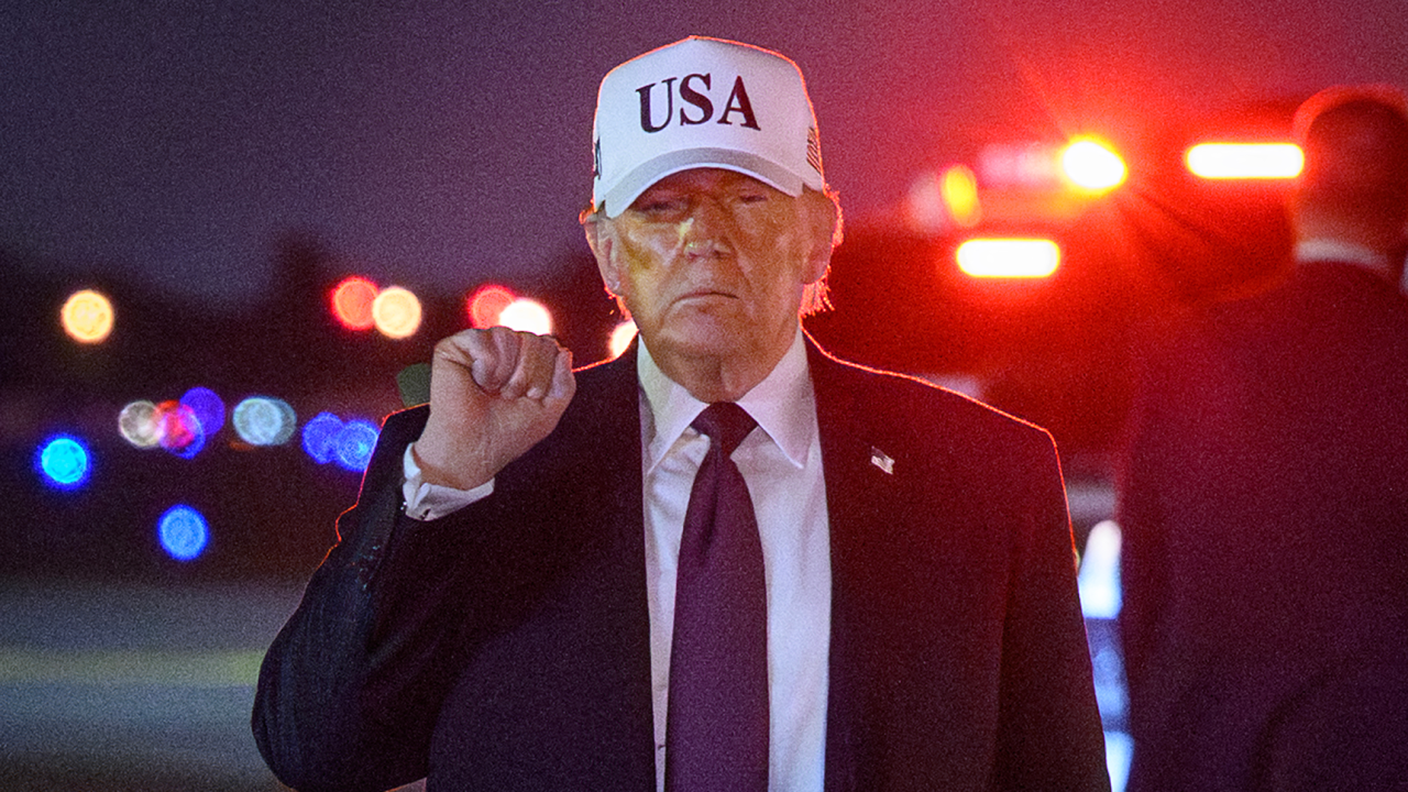 
                    Close up of Donald Trump with his fist in the air after landing at Palm Beach international airport. It is night time and he is wearing a white cap with USA  on it.
     