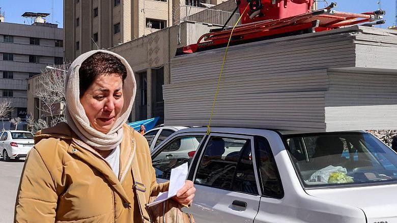 
                    A woman appears to be crying as she walks on a street in Tehran. She has a light coloured headscarf and a tan coat on. She is holding a piece of paper in her hand and has