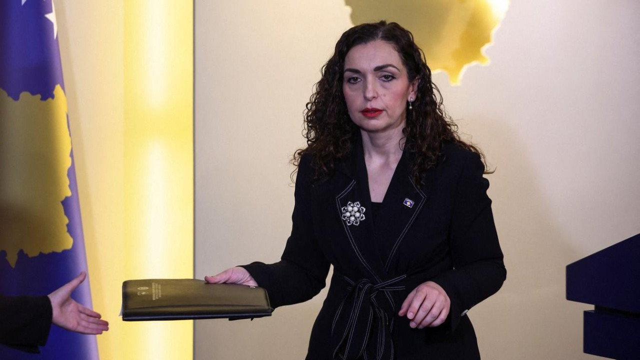 Vojsa Osmani gleda u pod u desnoj ruci drži svesku, a desno od nje je zastava Kosova