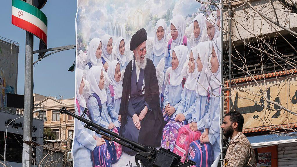Mušarac u vojničkoj uniformi drži oružje u ruci iza njega plakat ubijenih devojčica i iranskog vođe