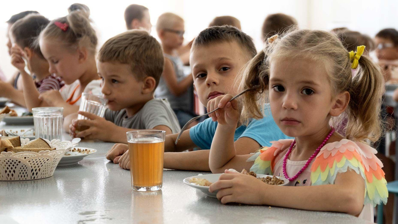 Deca iz sirotišta u Donjeckoj oblasti jedu obrok u kampu u Zolotoj Kosi, naselju na Azovskom moru, Rostovska oblast, jugozapadna Rusija, petak, 8. jul 2022. Devojčica sa pletenicama je desno, najbliža kameri, tužno gleda u kameru i drži viljušku u desnoj ruci iznad tanjira hrane. Čaša crveno-smeđe tečnosti je na stolu pored nje. Dečak pored nje takođe gleda u kameru. Ostala deca su zauzeta jedući i pijući.