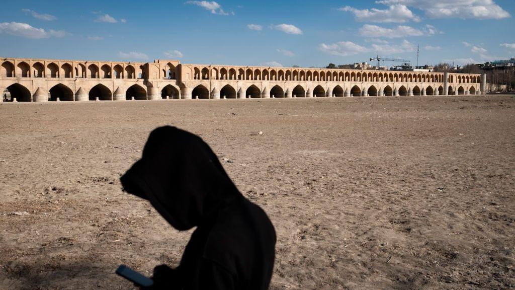 Iranac koristi mobilni telefon dok stoji na osušenoj obali reke Zajandeh Rud, sa istorijskim mostom Si-o-se-pol (33 mosta) vidljivim u pozadini, u istorijskom gradu Isfahanu, Iran, početkom 2025. godine.