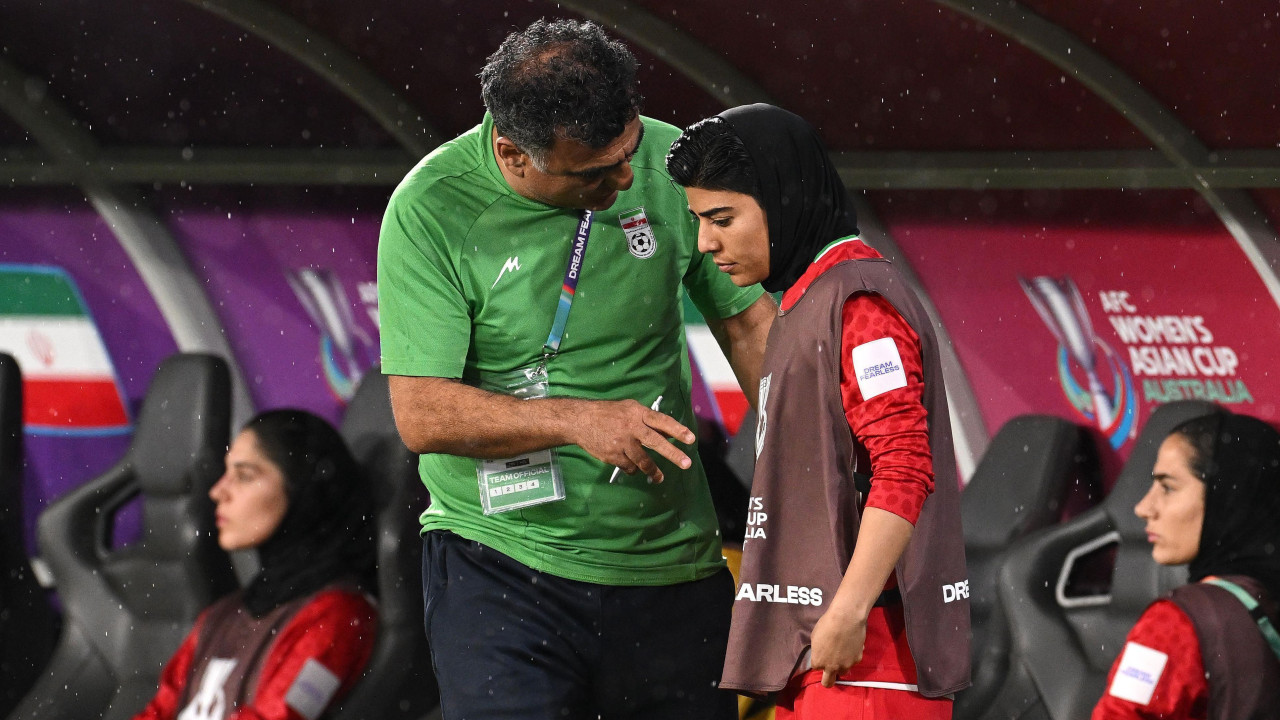 Iranski trener obučen u zeleno razgovara sa igračicom Monom Hamudi, koja nosi crvenu majicu sa smeđom majicom bez rukava, tokom utakmice, dok još dve fudbalerke sede sa svake strane. 