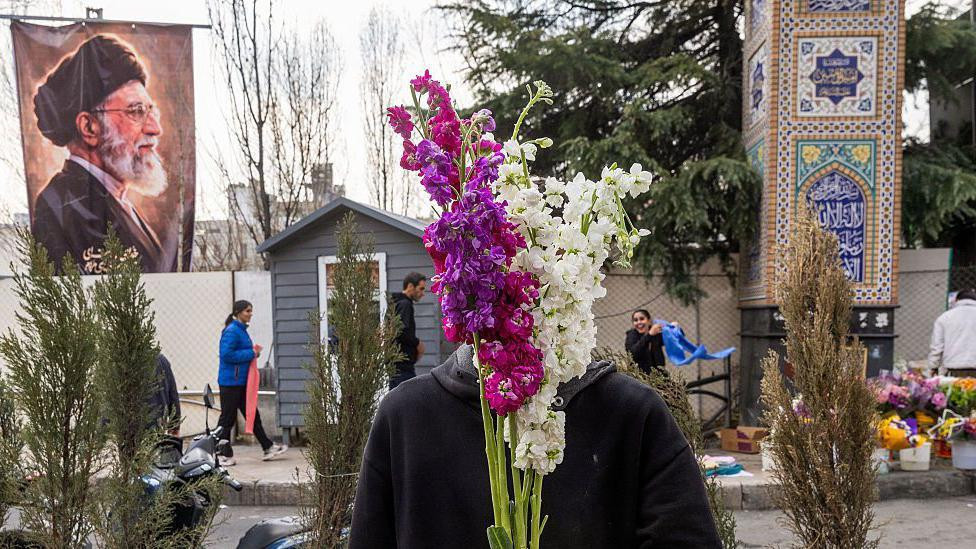 
                    A flower vendor holds purple and white flowers over his face as people shop in preparation for Nowruz celebrations on 19 March 2026 in Tehran, Iran. Behind him on the lef