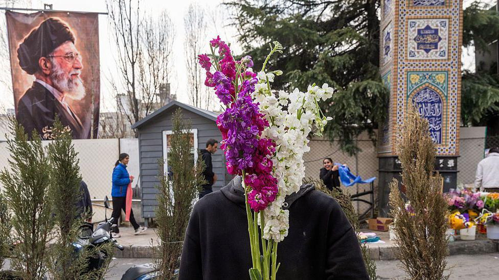
                    A flower vendor holds purple and white flowers over his face as people shop in preparation for Nowruz celebrations on 19 March 2026 in Tehran, Iran. Behind him on the lef