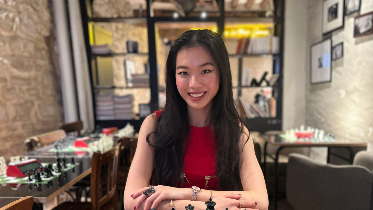 A young woman sitting in front of a chess board in an empty bar. She is wearing a red dress, her hair is dark long and straight. She is smiling at the camera and her hands are on the table before the board 