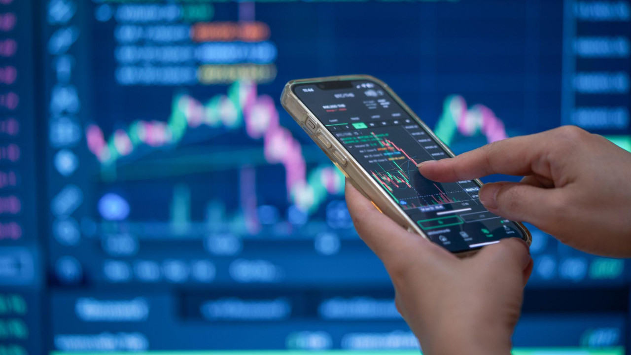 A trader's hands using their phone to make a trade in front of a trading screen in soft focus