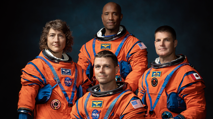 Na slici je četvoro astronauta u jarko narandžastim svemirskim odelima kako poziraju za formalni grupni portret na tamnoj pozadini. Odela su glomazna sa kaiševima i džepovima, što naglašava njihov tehnički dizajn. Troje astronauta stoje, a jedan sedi, svi okrenuti ka kameri. 