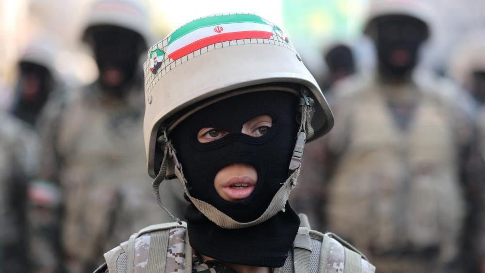 Mladi muškarac u uniformi i sa kacigom sa iranskom zastavom, iza njega su vojnici