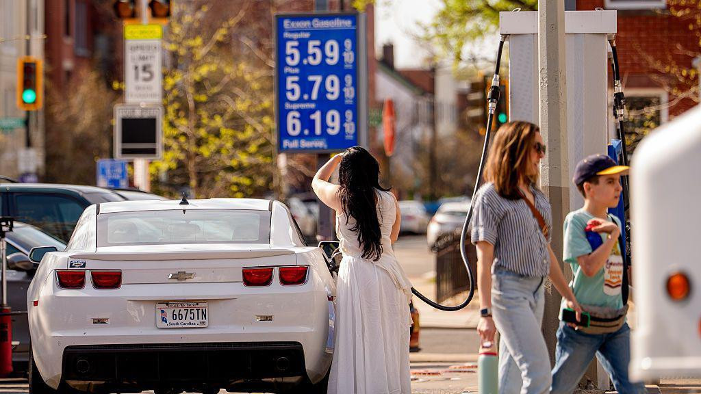 Žena u beloj haljini stoji u centru fotografije. Ona koristi pumpu za beli automobil na benzinskoj pumpi u Vašingtonu
