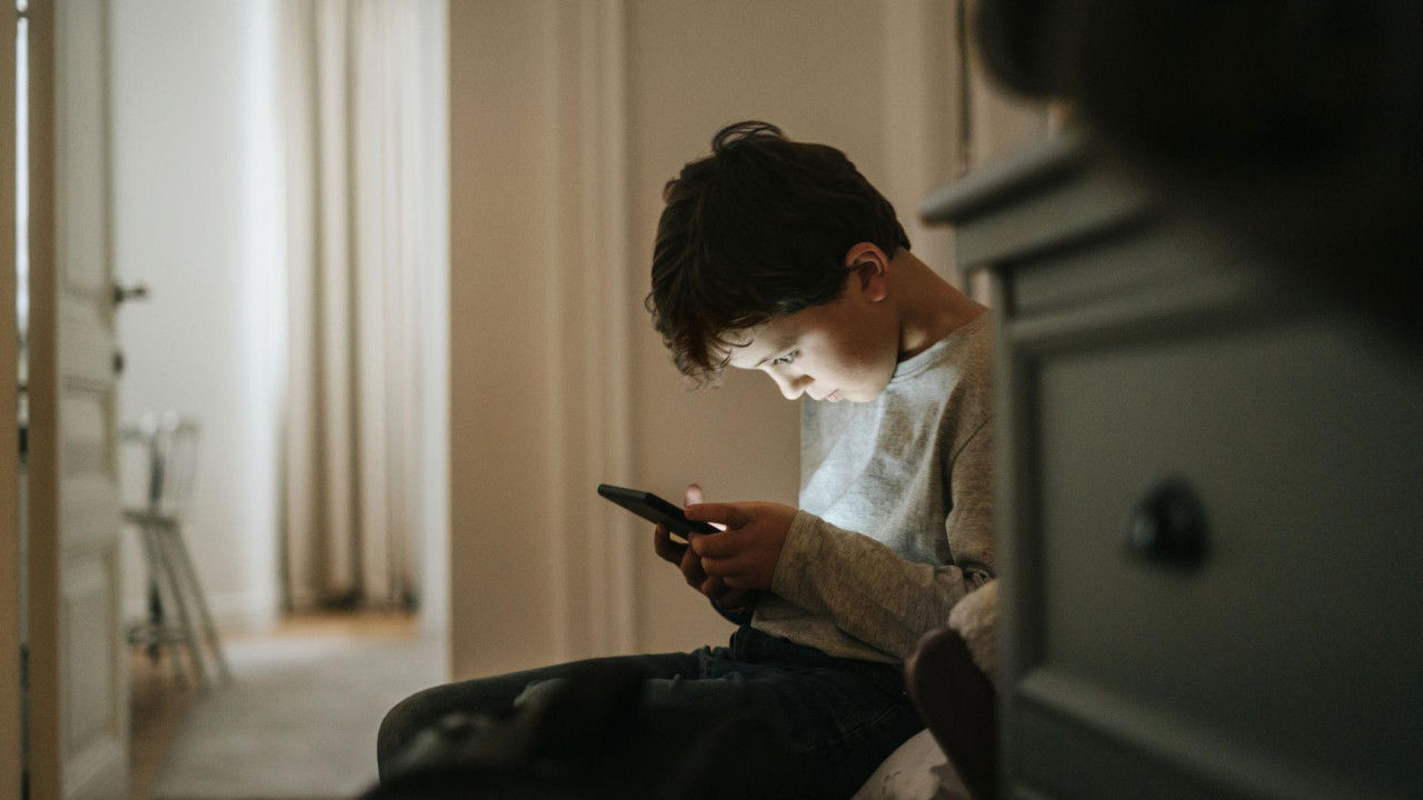 Dečak gleda u ekran mobilnog telefona, sedi na fotelji u sobi, iza komode