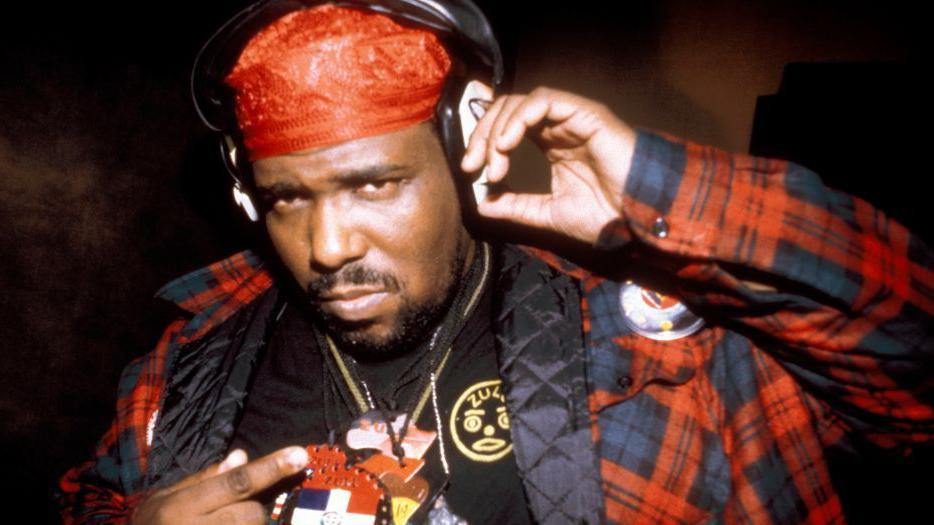 Afrika Bambata pozira sa slušalicama i crvenom maramom dok pokazuje na ambleme na ogrlicama i košulji