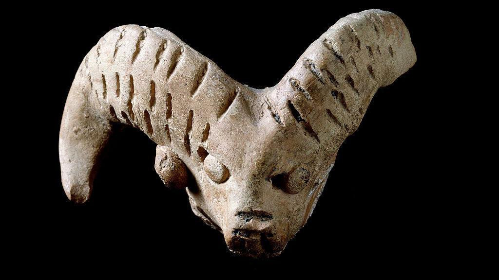 A statue in beige rock of a goat head, with rows of short lines engraved on its horns, against a black background. One of the horns appears to be missing a piece at the end.