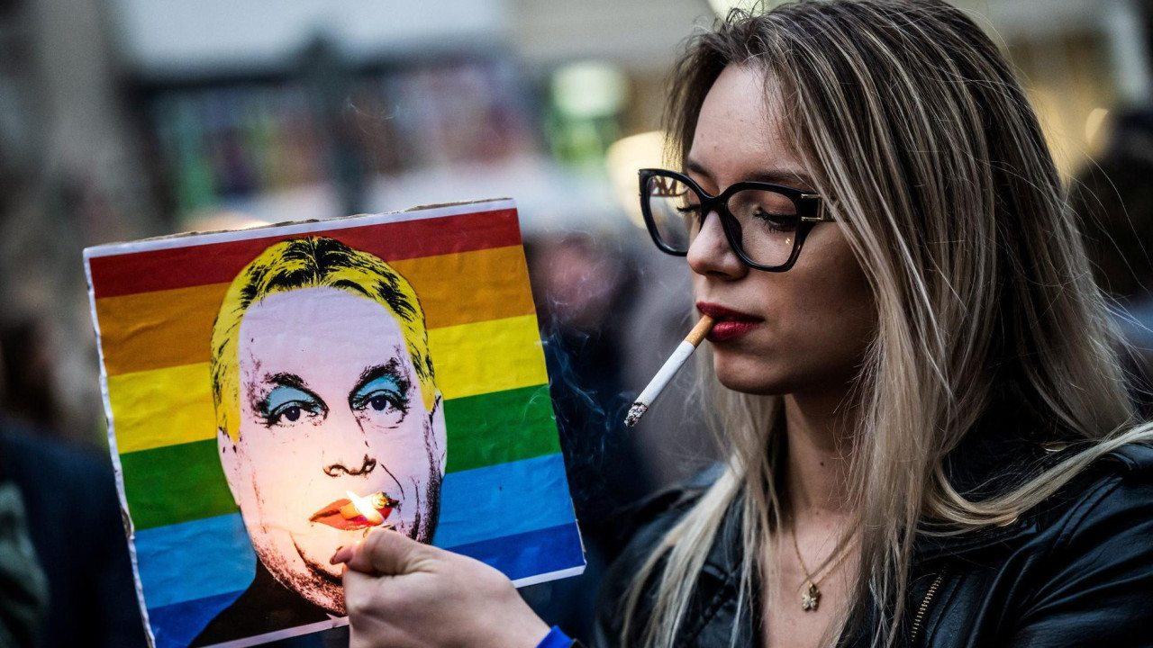 Devojka sa naočarima za sunce i cigarom u ustima u levoj ruci drži upaljač, a u desnoj poster sa Orbanovom slikom dok je pozadina zastava LGBT zajednice