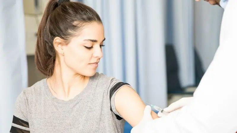 Devojka gleda u levu ruku dok kojoj medicinski radnik daje vakcinu