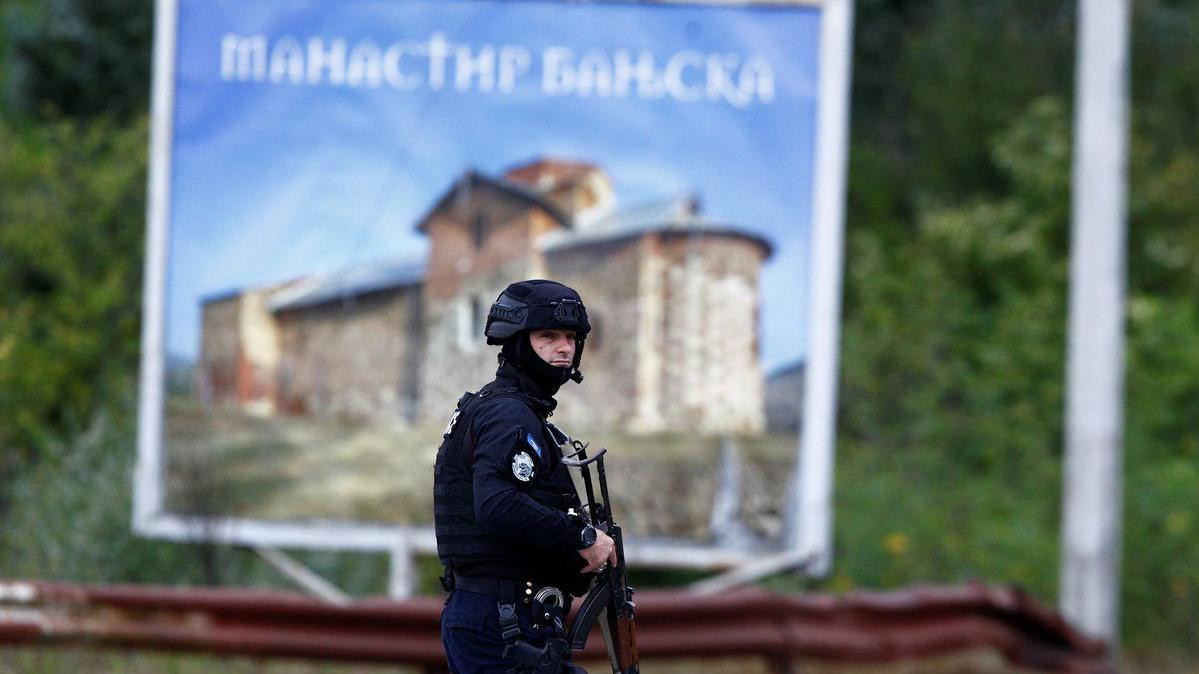 Policajac u kosovskoj uniformi ispred bilbodra na kojem je slika manastira i ćirilični naziv Manastir Banjska