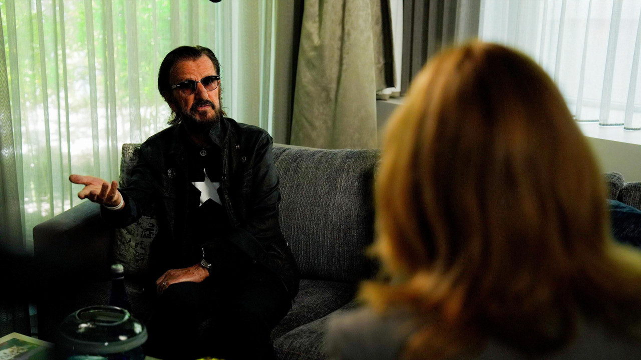 Ringo Star u prvom planu sedi na kauču i razgovara sa ženom okrenutom leđima ka kameri