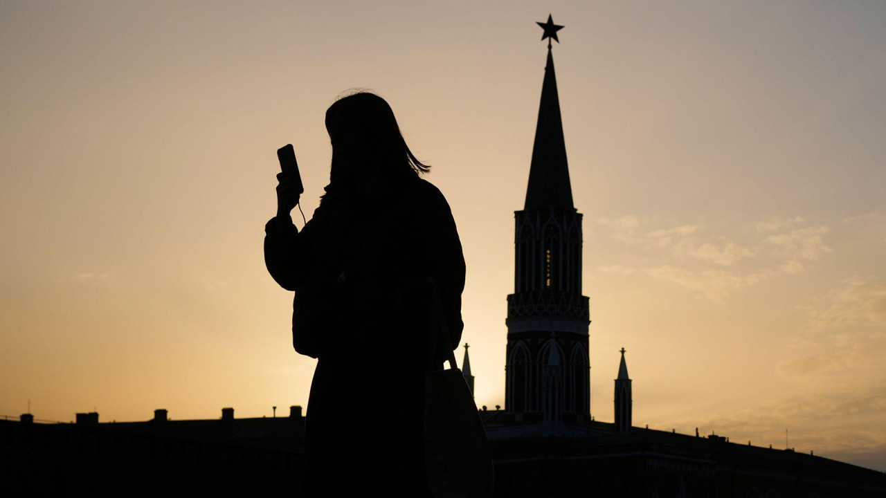 
                    A person is silhouetted against a glowing orange sky as they look at their smartphone, with the silhouette of the Kremlin in the background during sunset in the Red Squar