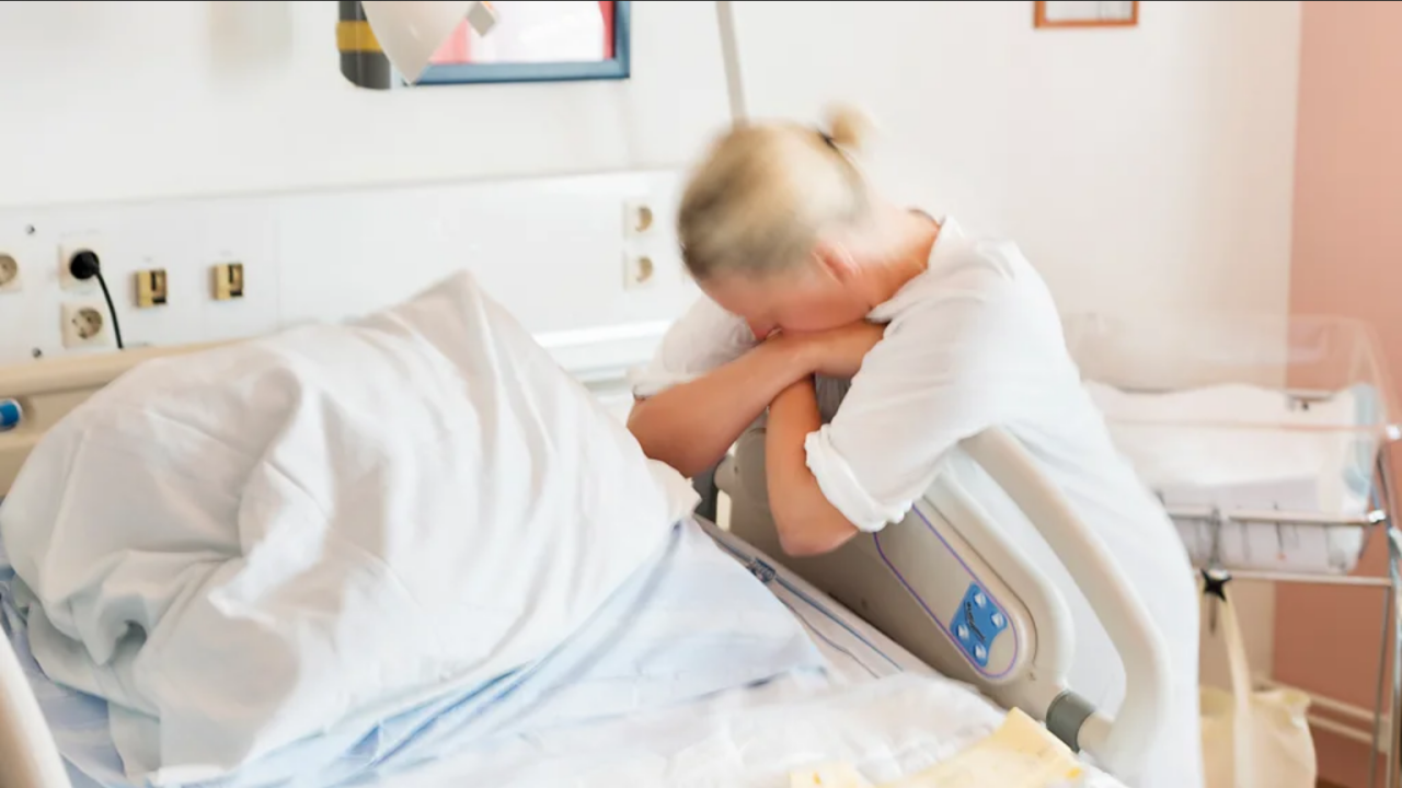 Žena pored bolničkog kreveta na nogama, grli dršku od kreveta, glava joj je na rukama