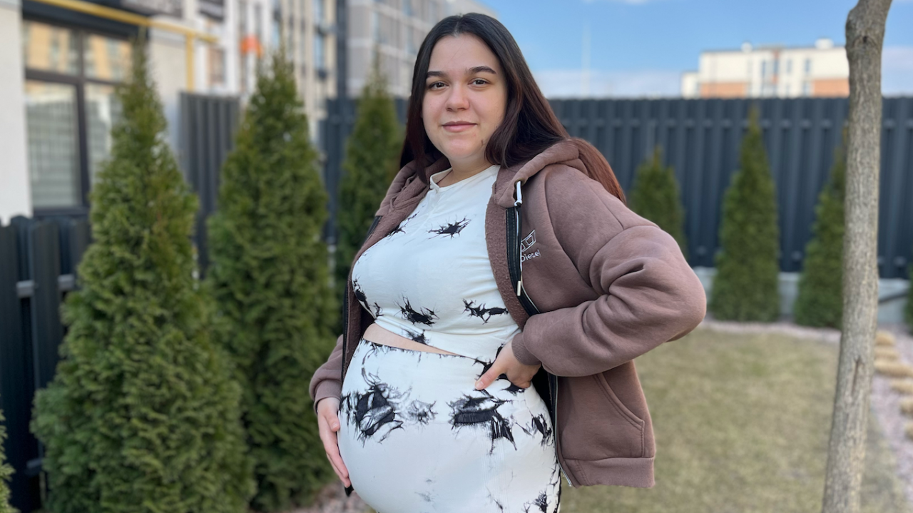 
                    Trudna žena stoji u bašti sa rukom na stomaku. Ima dugu, ravnu, tamnu kosu i nosi odgovarajući top i pantalone koje su bele sa nekoliko crnih mrlja. Takođe ima svetlo sm