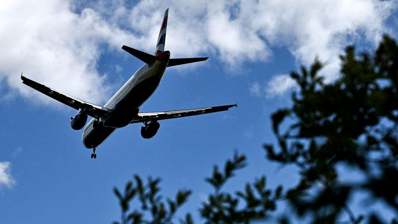 A silhouetted plane flies overhead with shrubbery in view against a blue sunny-cloudy sky while arriving at Gatwick Airport on 20 April.