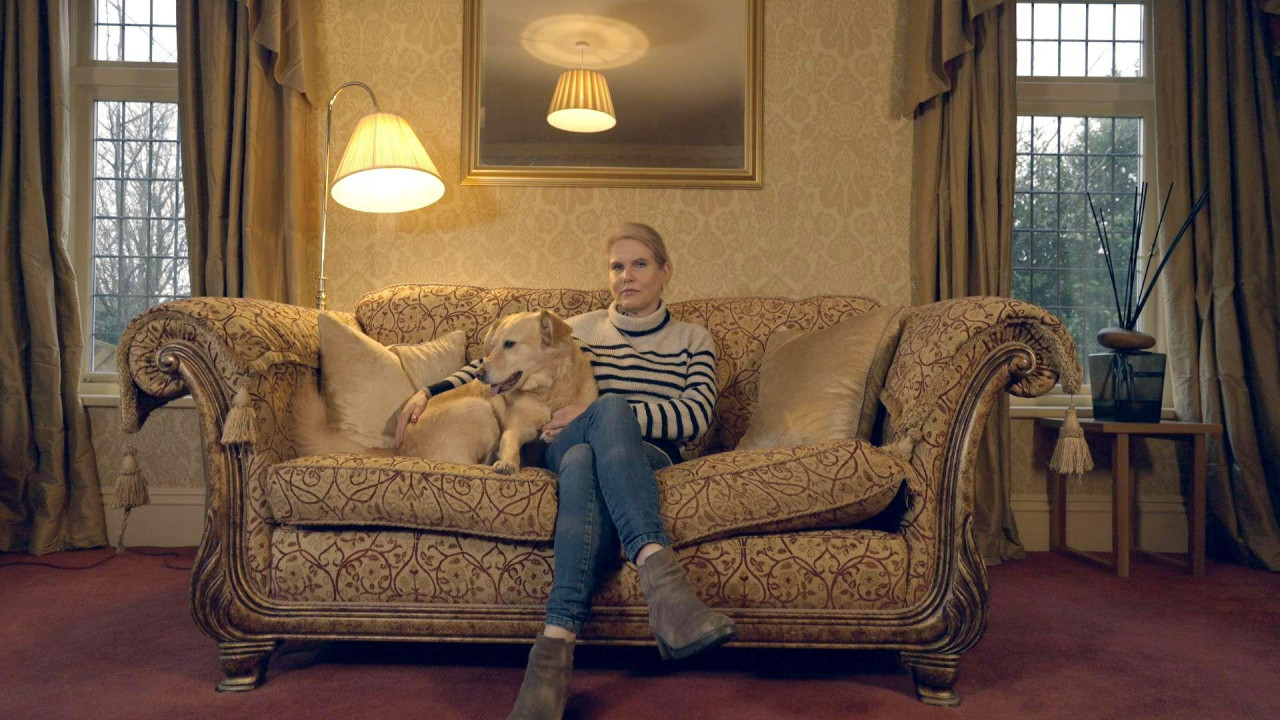 Nikola Berd u džinsu, kratkim čizmama i prugastom džemperu sedi na šarenoj sofi prekrštenih nogu. Desnom rukom mazi psa. Ona sedi u dnevnoj sobi sa crvenim tepihom i otvorenim teškim zavesama na dva prozora sa obe stra 