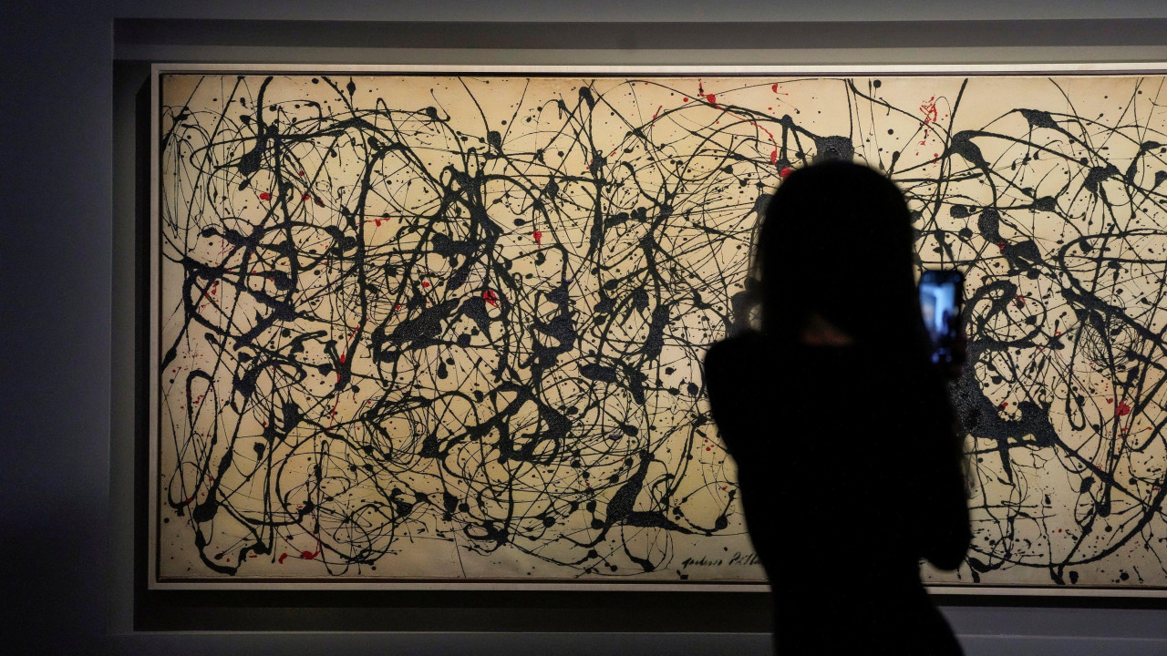 
                    A person with long hair in silhouette is seen facing the artwork holding their mobile phone to video the painting. The artwork is long horizontally and the grey canvass a