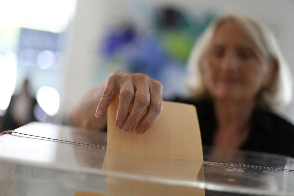 žena ubacuje žuti listić u glasačku kutiju