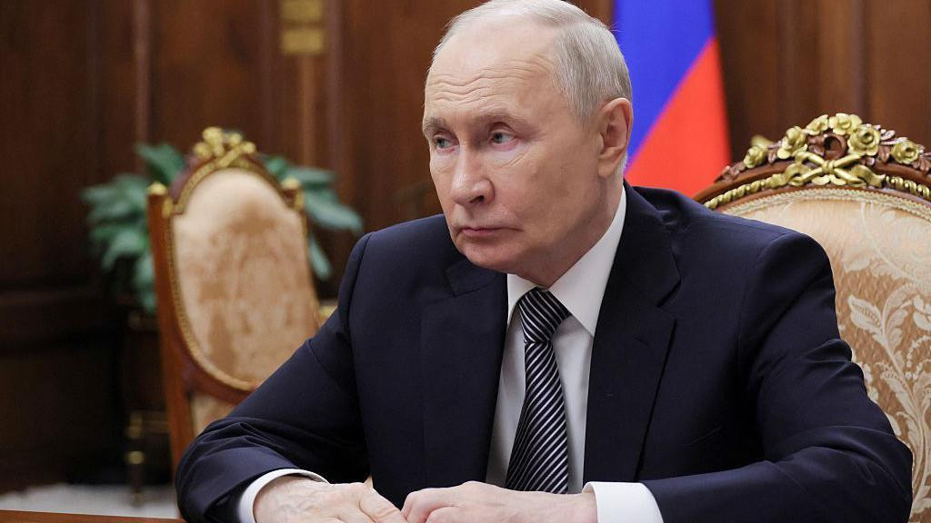 Vladimir Putin u teget odelu za stolom gleda u stranu, a iza njega se nazira ruska zastava