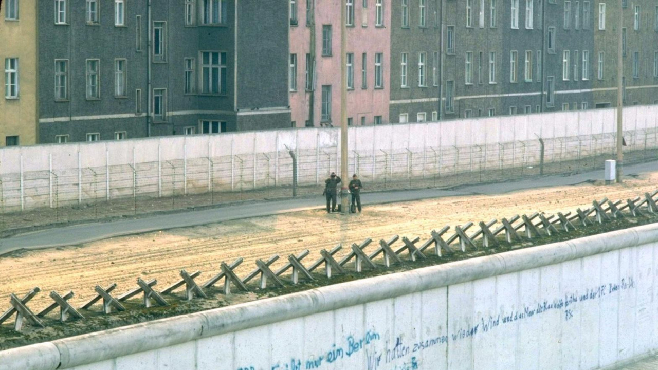 Beli betonski Berlinski zid 1. marta 1982. godine, sa ispisanim grafitima. Sa druge strane zida je deo neplodne zemlje sa dva istočnonemačka stražara, metalnom ogradom, još jednim belim betonskim zidom, a zatim zgradama u Istočnom Berlinu.