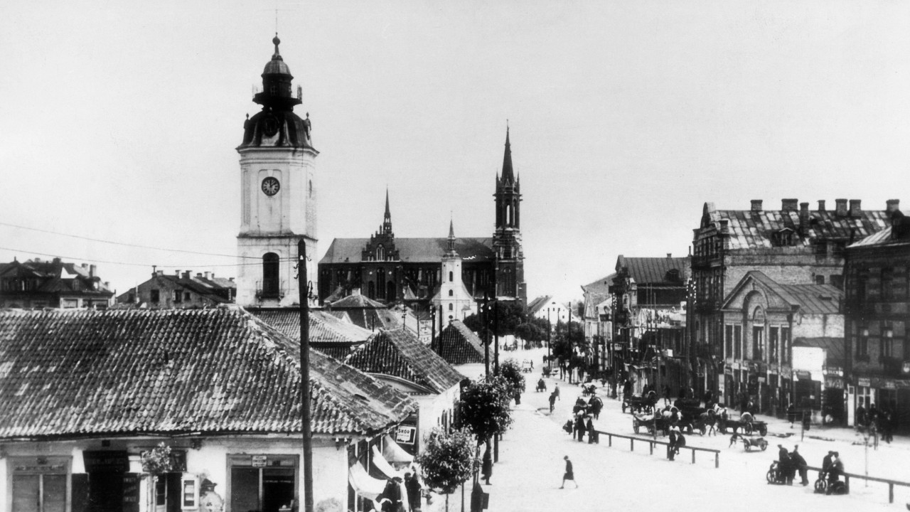Ugao ulice sa zgradom sa kosim krovom i tornjem. Nadstrešnice se protežu iznad trotoara sa jedne strane. Visoka zgrada sa tornjem je u daljini, a iza ograde sa druge strane ulice nalaze se zgrade različitih visina. Nekoliko ljudi hoda ulicom koja izgleda kao da je prekrivena snegom. 