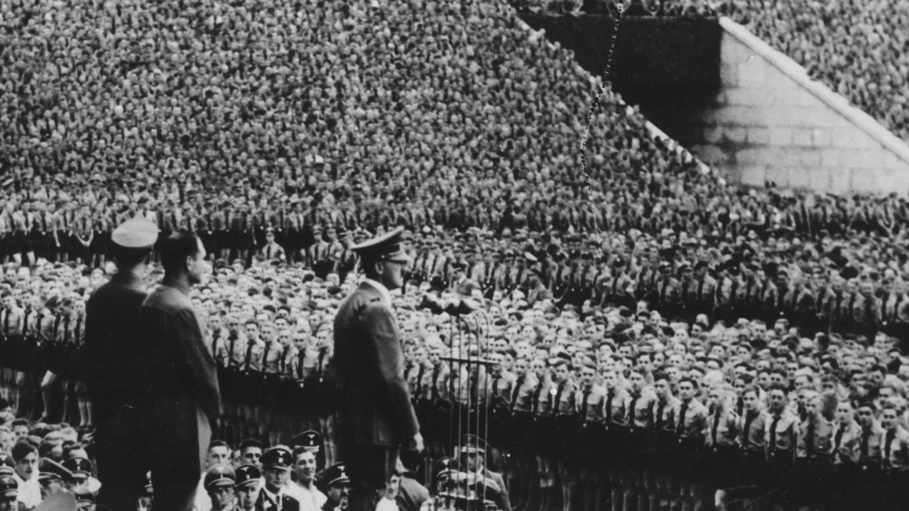 Adolf Hitler stoji pred hiljadama mladih ljudi na stadionu 12. septembra 1938. Iza njega su Baldur fon Širah, vođa Hitlerove omladine, i Rudolf Hes, Hitlerov zamenik