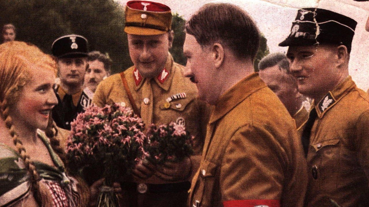 Žena sa plavom kosom spletenom sa strane gleda Hitlera dok joj razgovara okružen muškarcima u vojnim uniformama 